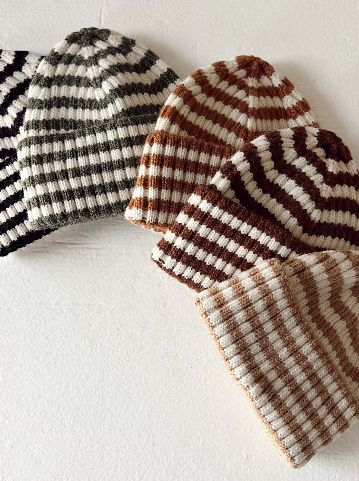 Five striped beanies in black, green, brown, and cream colors arranged on a light background.