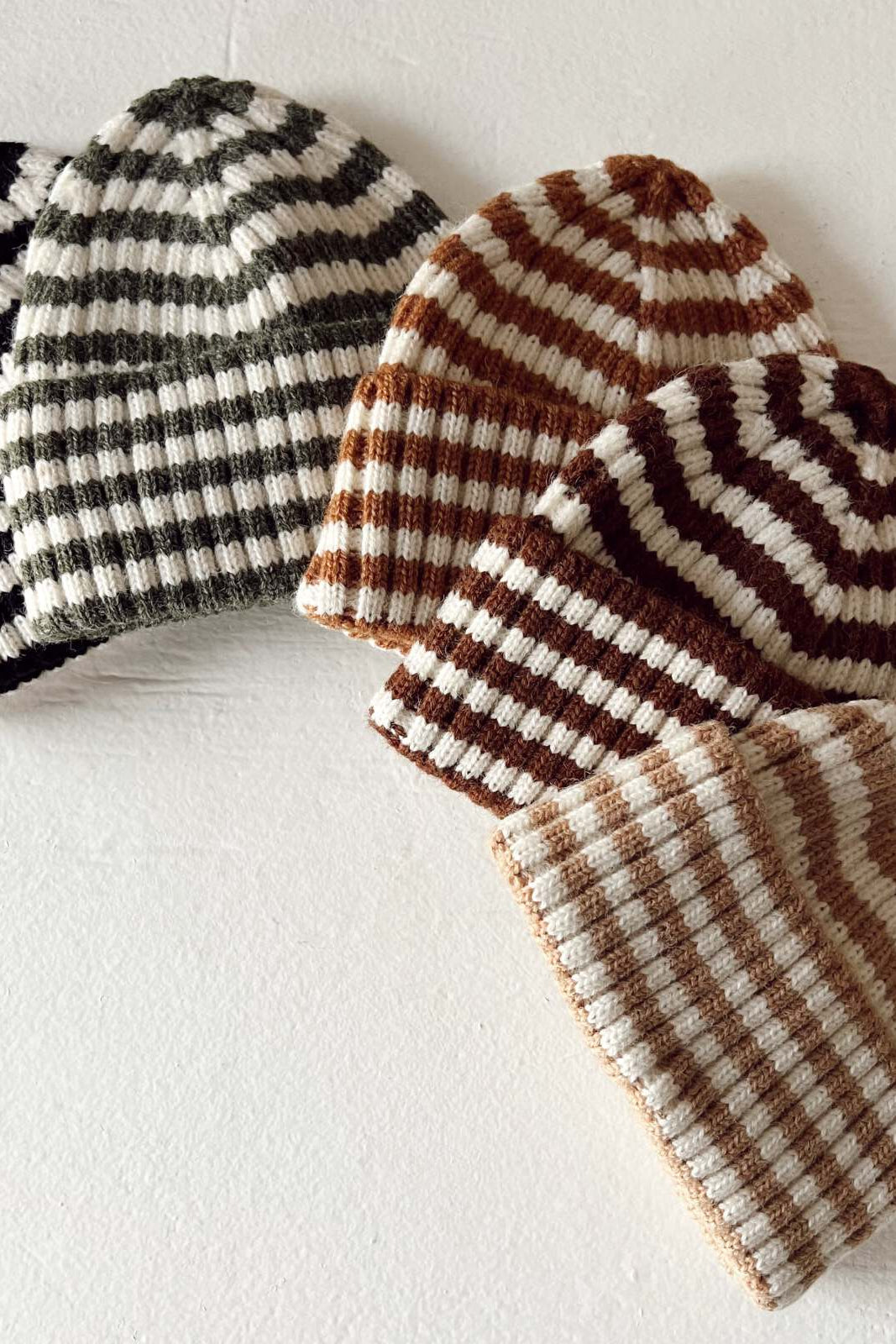 Five striped beanies in black, green, brown, and cream colors arranged on a light background.