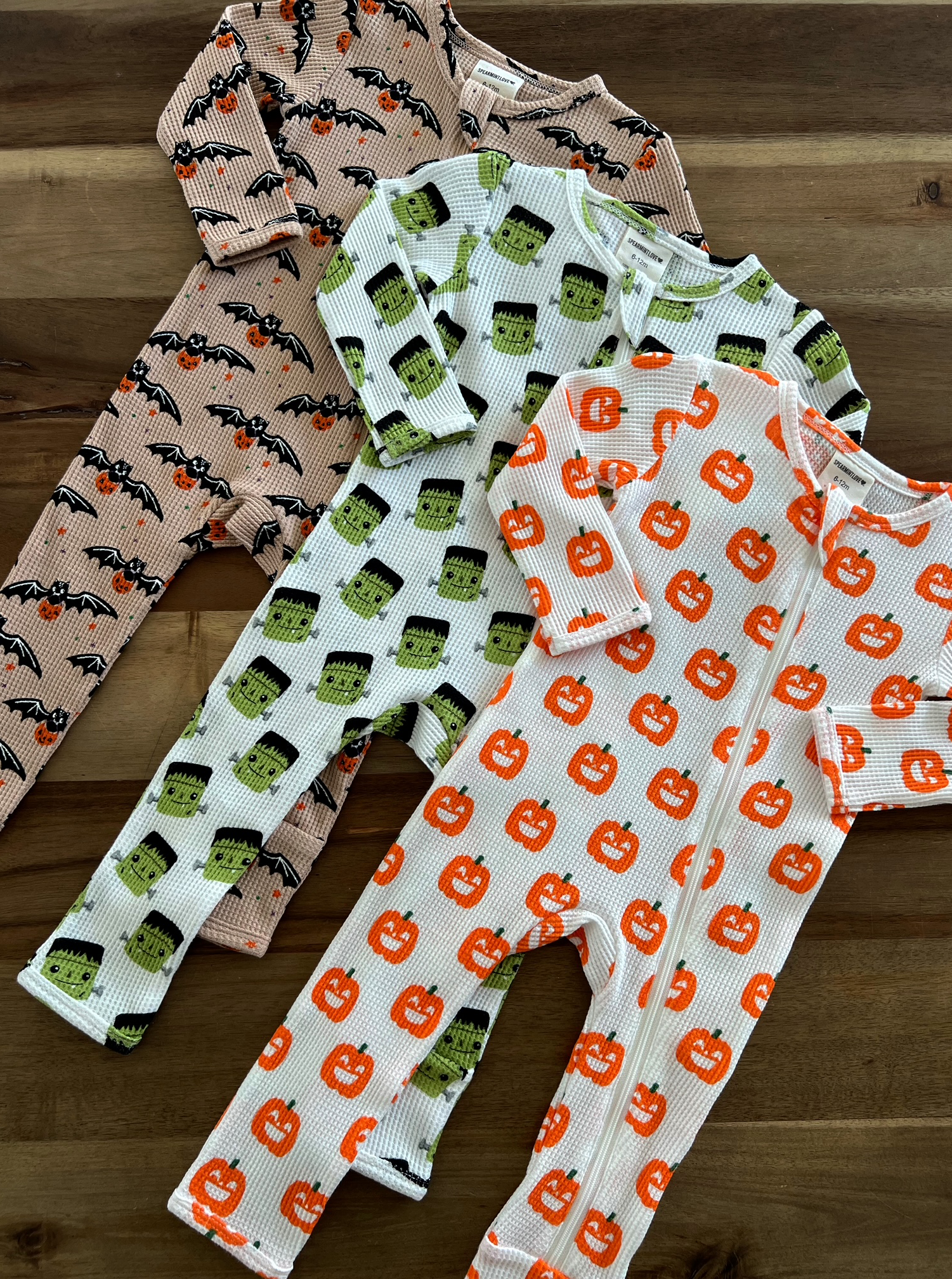 Three Halloween-themed baby pajamas featuring bats, Frankenstein, and pumpkins on a wooden surface.