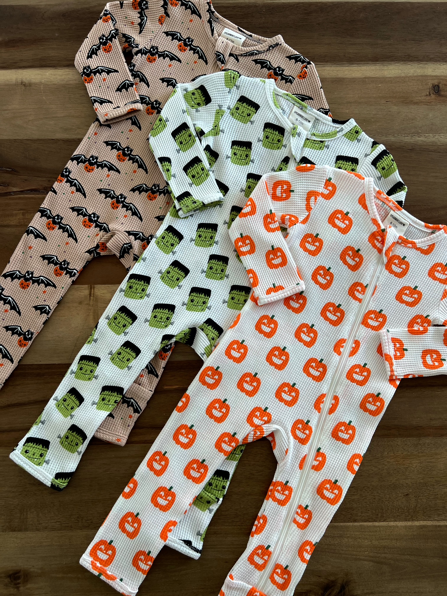 Three Halloween-themed baby pajamas featuring bats, Frankenstein, and pumpkins on a wooden surface.