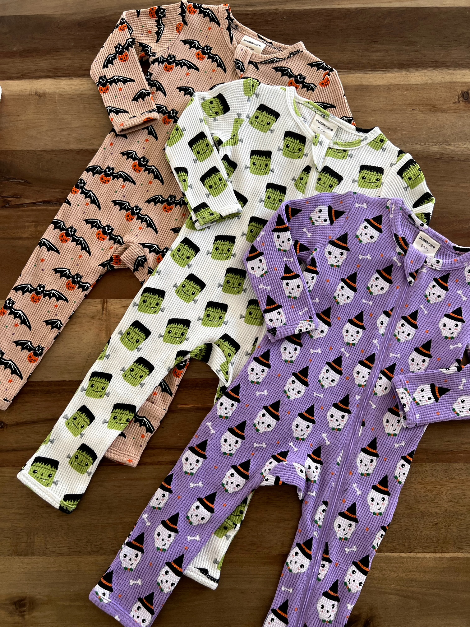 Three patterned pajamas featuring bats, Frankenstein, and skulls in Halloween themes on a wooden surface.