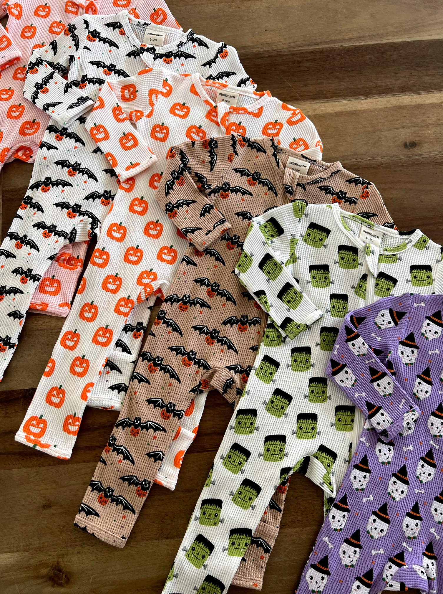 Multi-colored children's pajamas featuring Halloween designs like bats, pumpkins, and monsters arranged on a wooden surface.