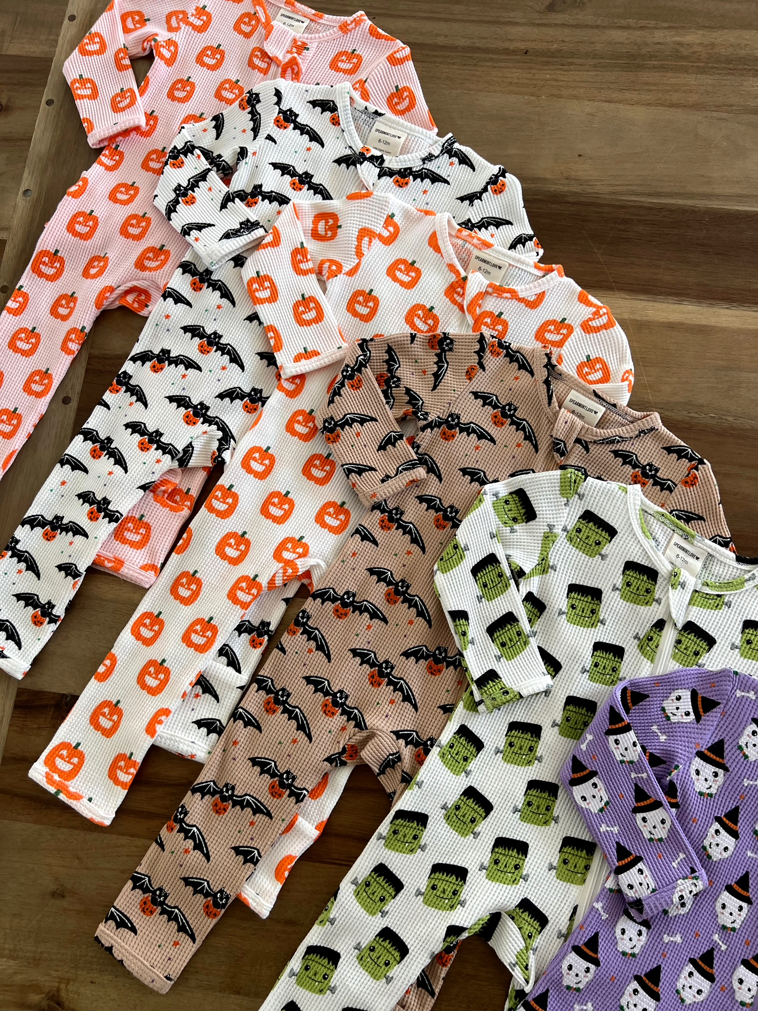 Colorful Halloween-themed baby pajamas featuring pumpkins, bats, and Frankenstein designs on a wooden surface.