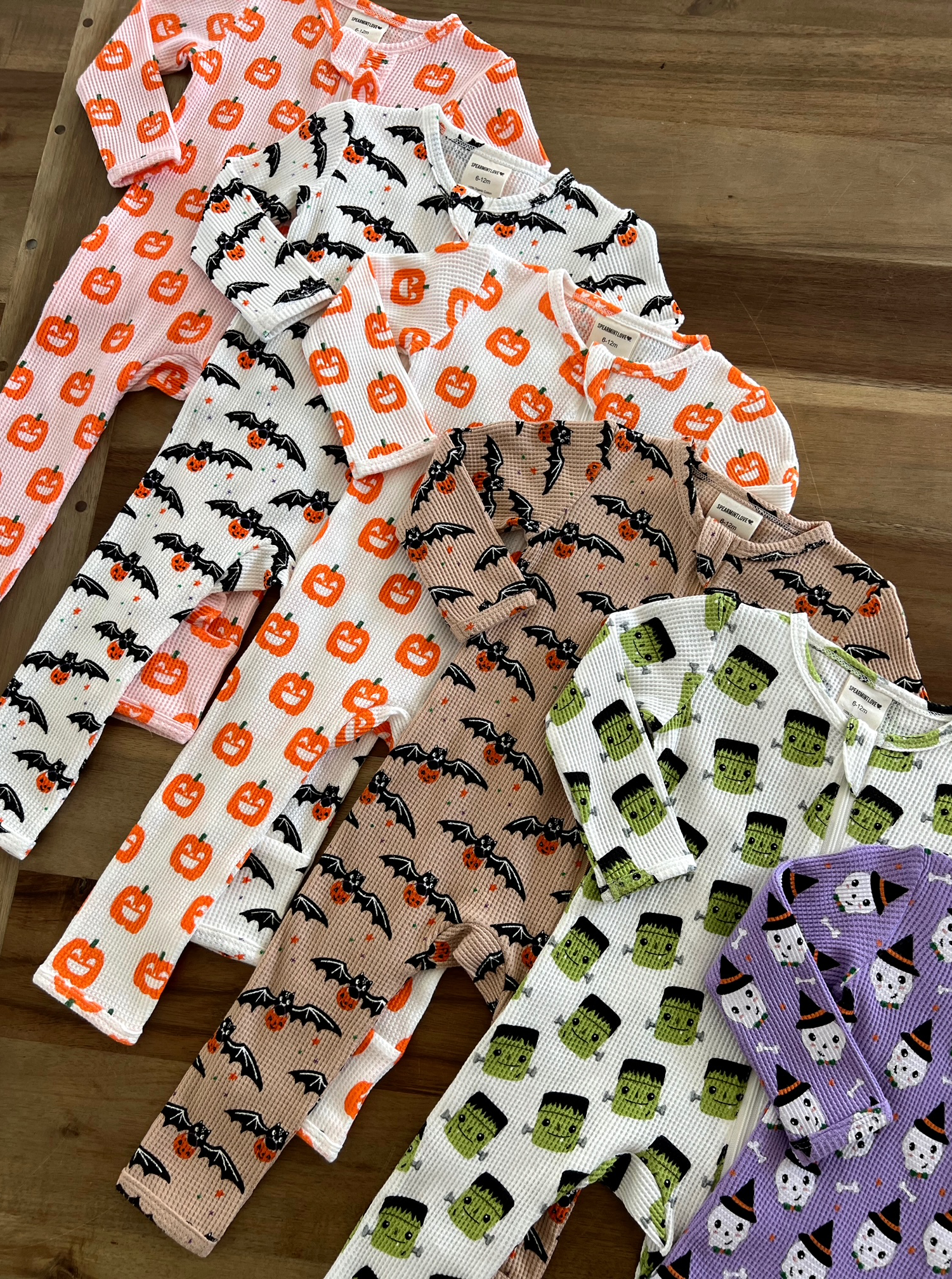 Colorful Halloween-themed baby pajamas arranged on a wooden surface featuring pumpkins, bats, and frankenstein prints.