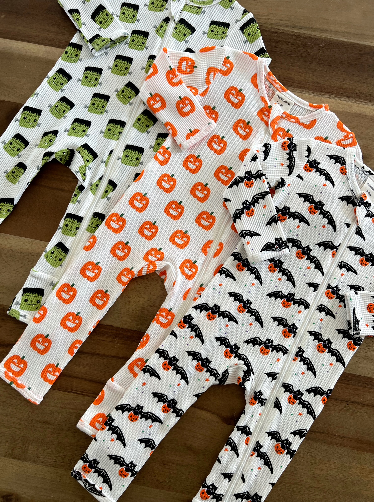 Three Halloween-themed onesies: green monster, orange pumpkins, and bats with pumpkins on a wooden surface.