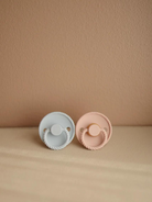 Two pacifiers, one in soft blue and the other in gentle pink, resting on a beige background.