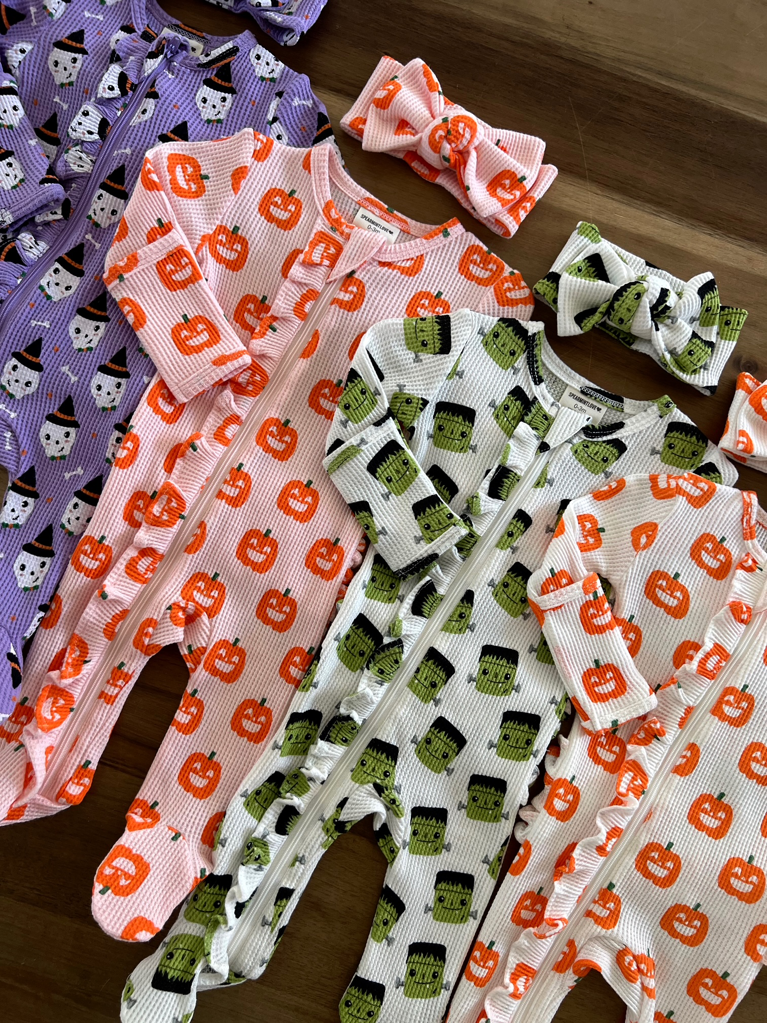 Colorful Halloween-themed infant rompers and headbands with pumpkins and monsters on a wooden surface.
