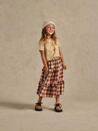 Smiling girl wearing a light shirt, plaid skirt, and beanie, standing against a neutral background.