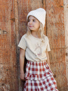 Young girl in a white beanie smiles, wearing a graphic tee and a red checkered skirt against a rustic wooden background.