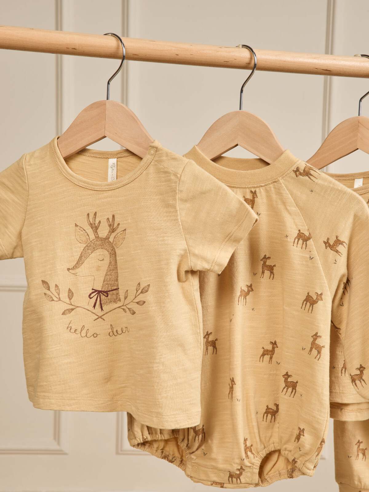 Tan baby clothes featuring a deer design and “hello deer” text, hanging on a wooden rack.