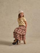 Smiling child in a cream shirt and plaid skirt, wearing a knit hat, sitting on a wooden stool.