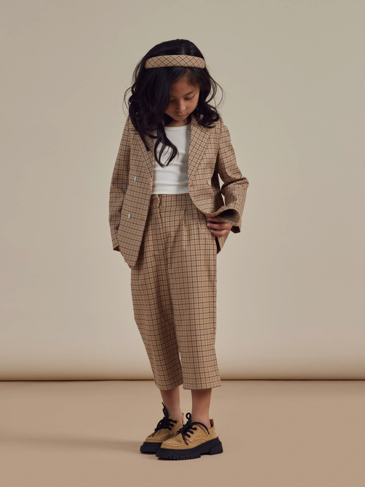 Young girl in a checked blazer and pants, wearing a white top, headband, and black sneakers, looking down.