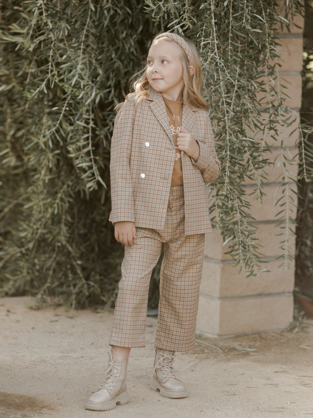 Young girl in a plaid suit and boots, posing outdoors near greenery, with a playful expression.