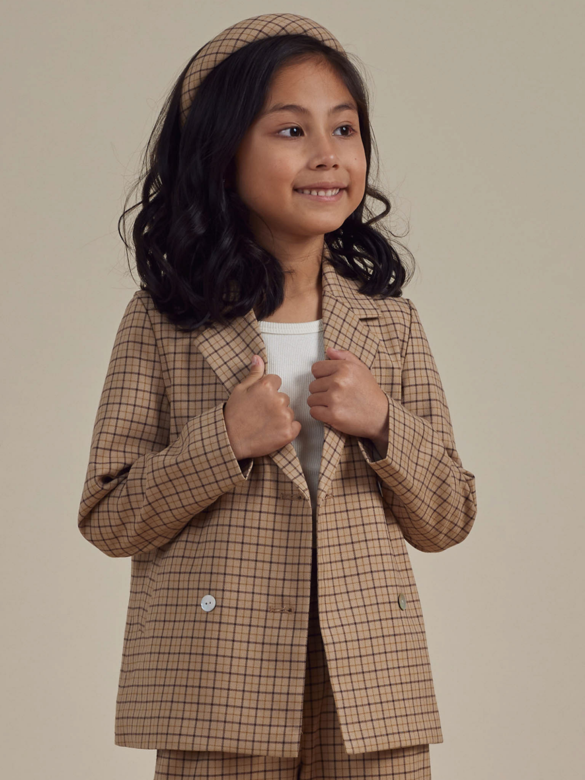 Young girl smiling in a plaid blazer and matching headband, posing against a neutral background.