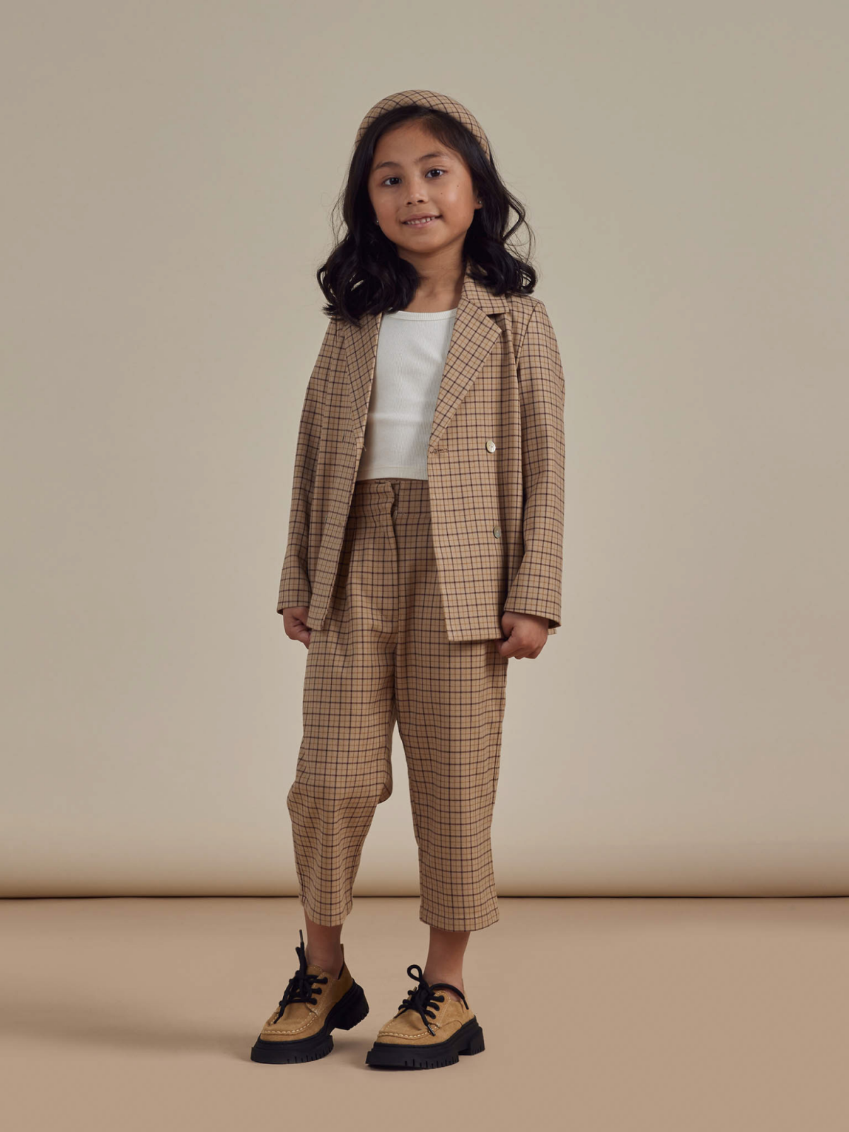 Smiling child in a beige plaid suit with a white top and black shoes, standing against a neutral background.