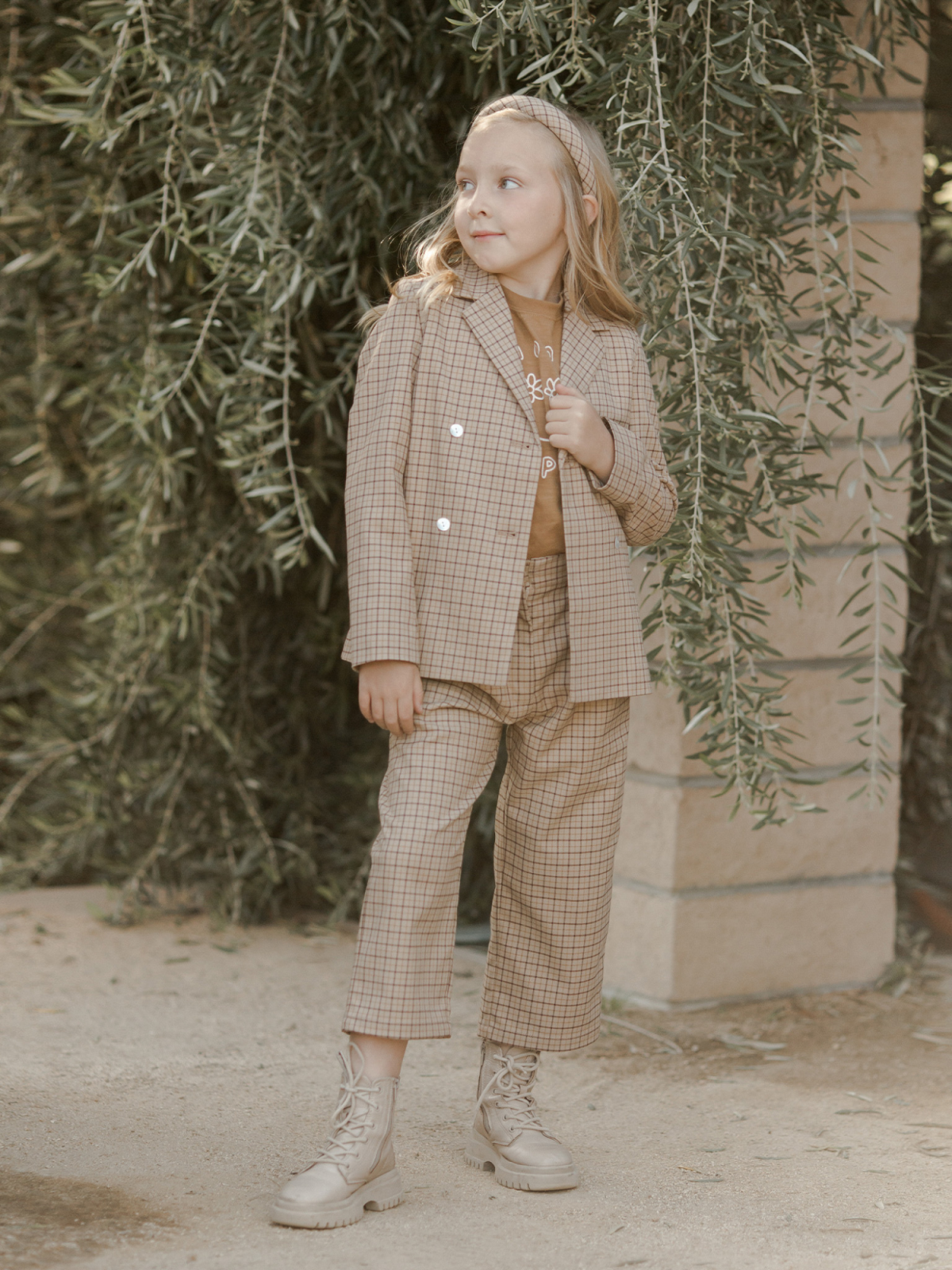 Girl in a stylish plaid suit and boots standing by greenery, posing thoughtfully in a natural setting.