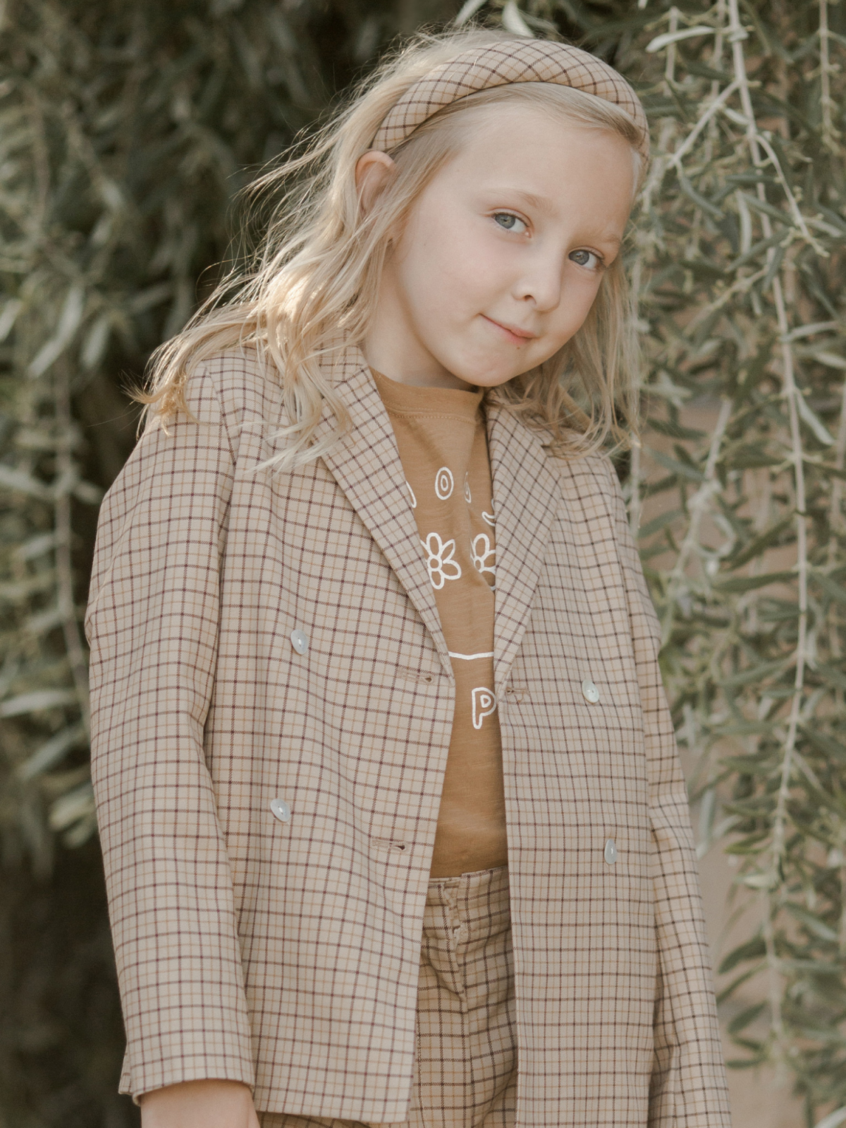 Blonde girl in a tan plaid suit jacket and headband posing outdoors among green foliage.