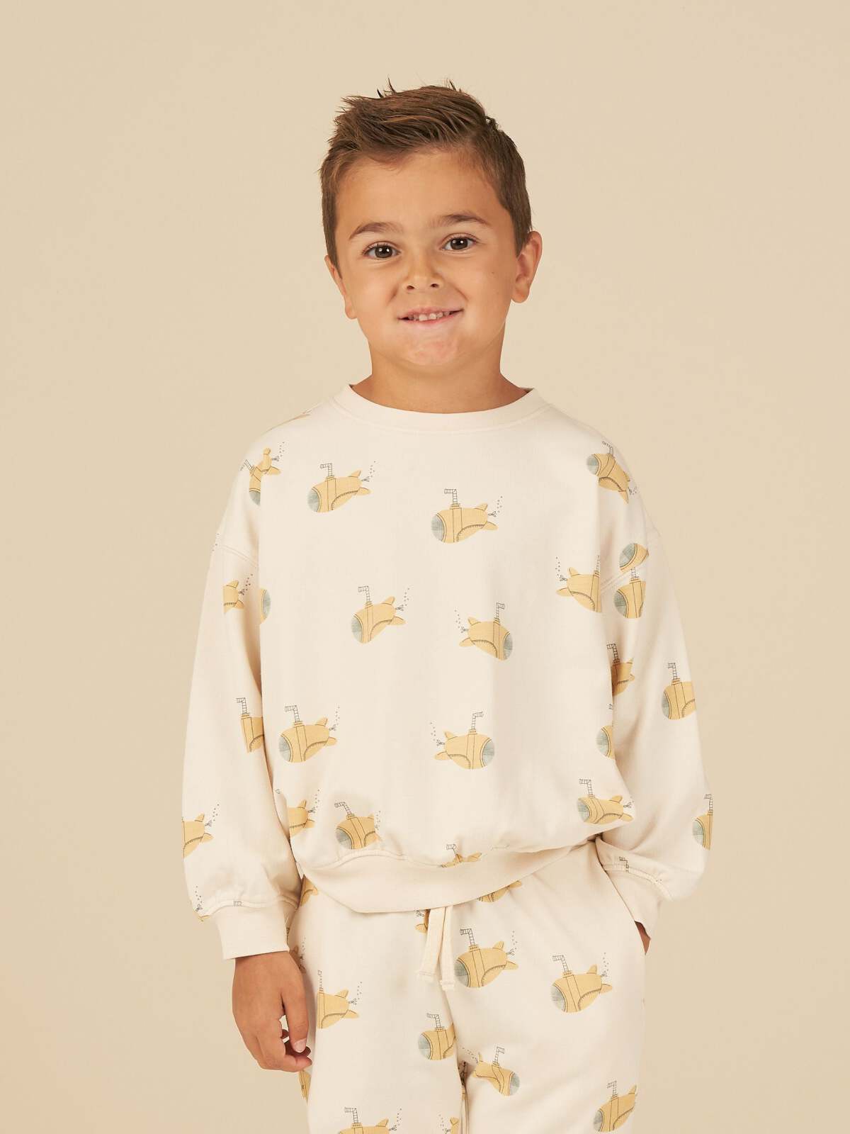 Smiling boy wearing a cream-colored sweatshirt and pants with submarine prints against a beige background.
