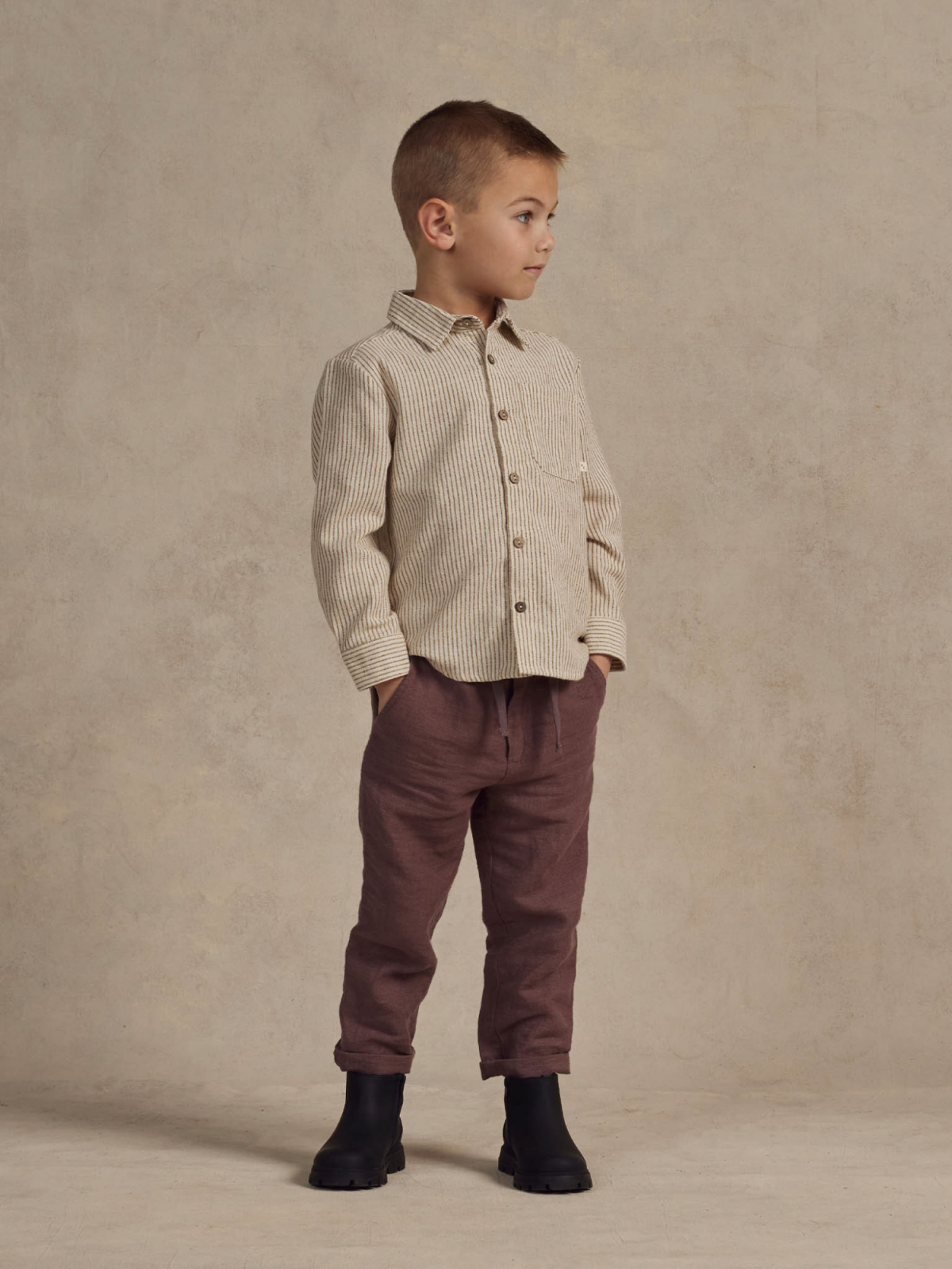 Child in striped shirt and burgundy pants stands confidently against a neutral backdrop.