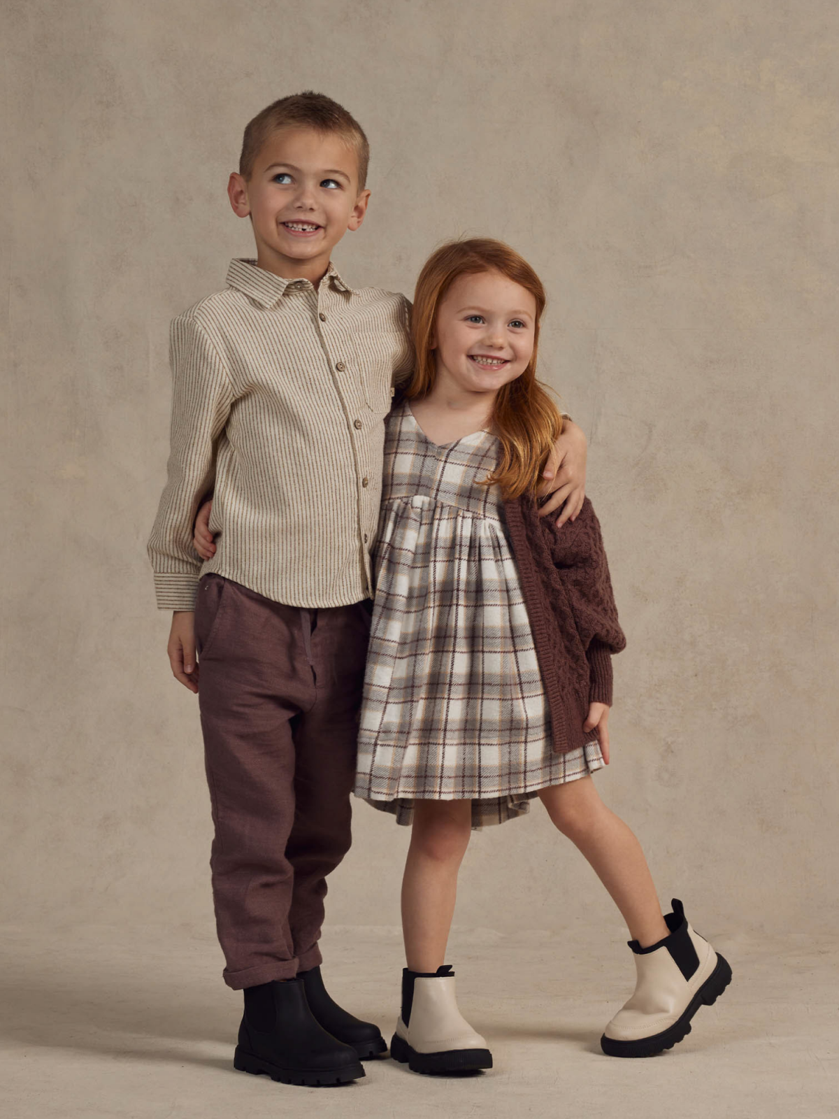 Two smiling children pose together, wearing cozy outfits and contrasting boots on a neutral background.