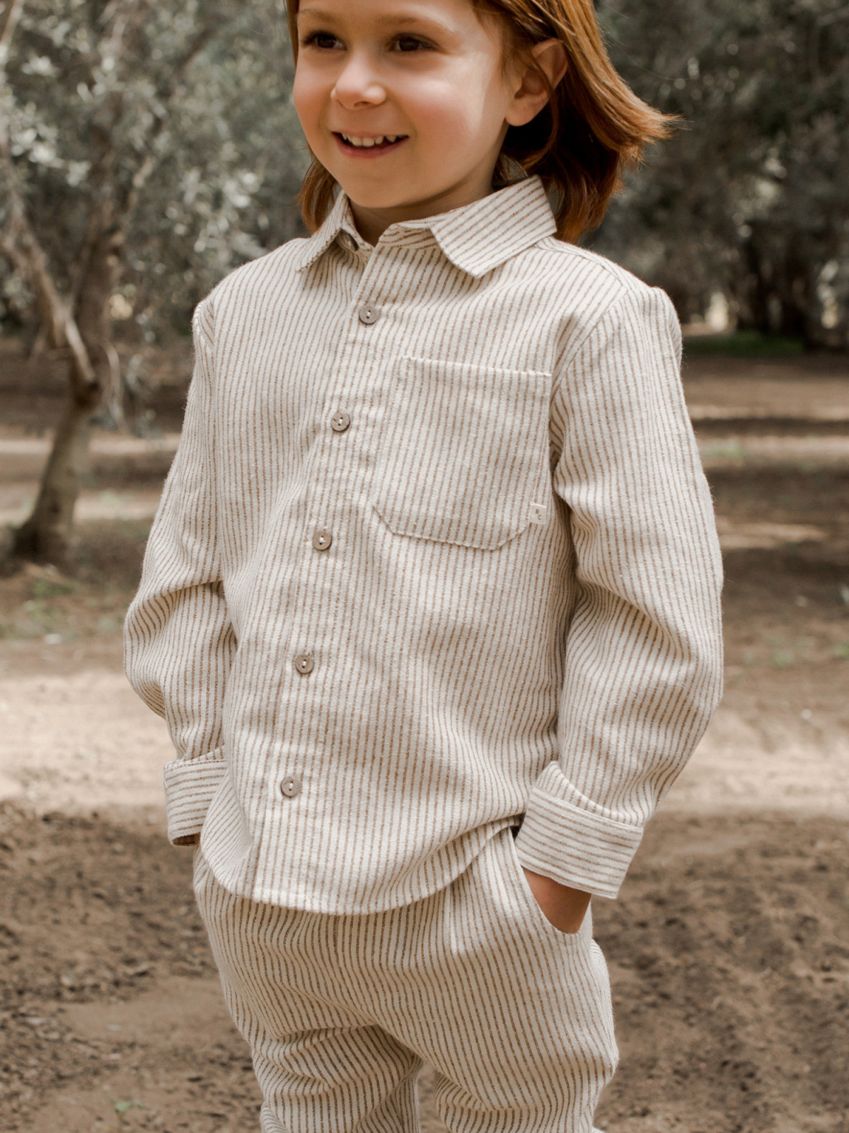 Smiling child in striped outfit stands outdoors, surrounded by trees, exuding joy and confidence.