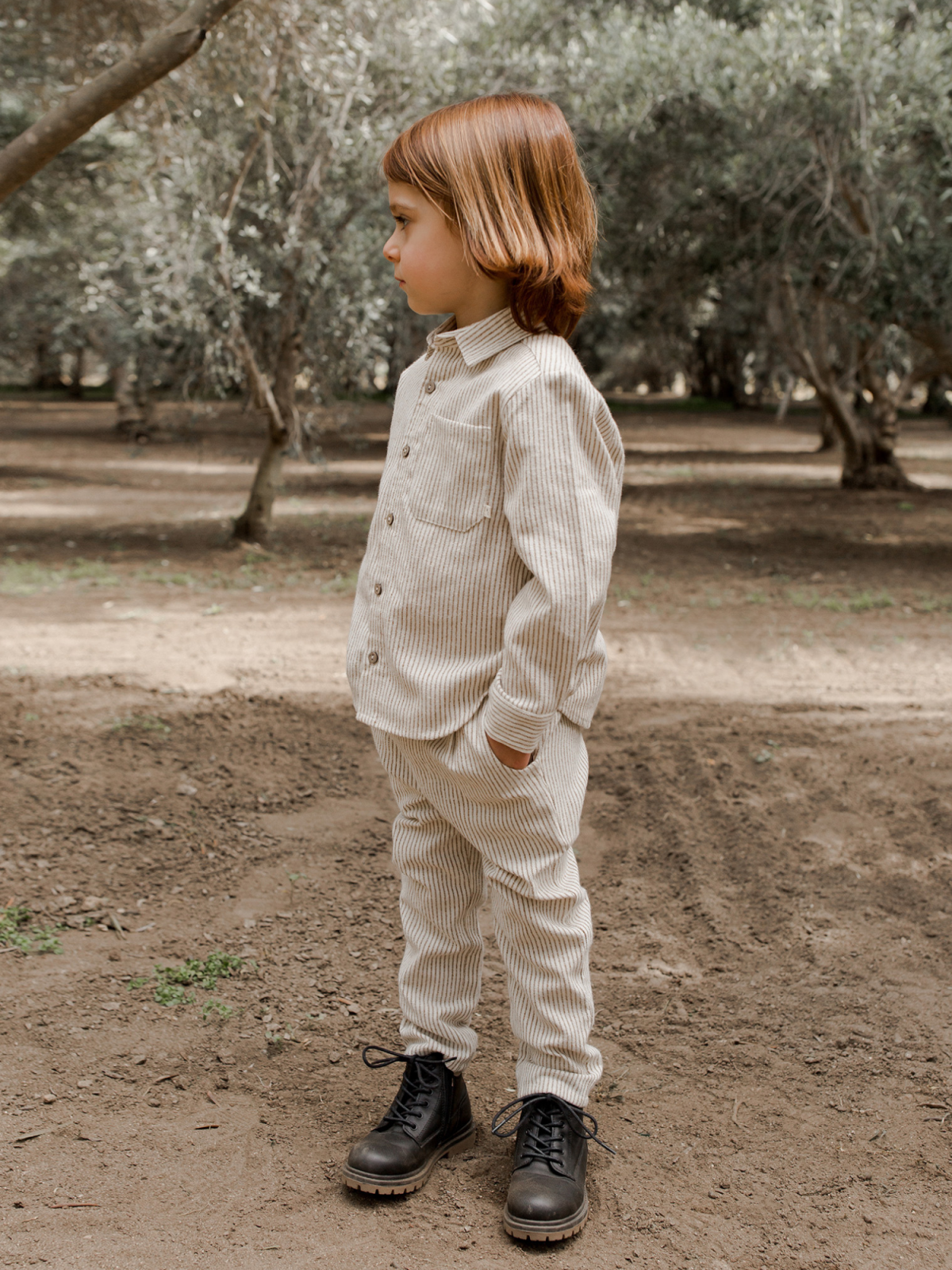 Child with shoulder-length hair in striped outfit and boots stands in an outdoor setting surrounded by trees.