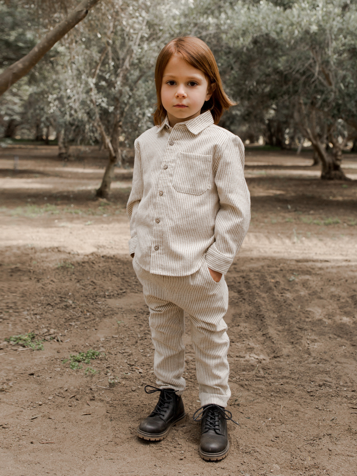 Child in a striped two-piece outfit stands in an outdoor setting surrounded by trees.