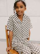 Child sitting on a wooden chair, wearing black and white checkered pajamas and a matching headband.