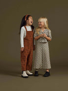 Two girls laughing while wearing stylish outfits against a muted background. One in an orange jumpsuit, the other in patterned attire.