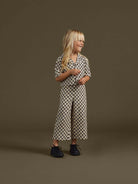 Smiling child in a checkered romper stands against a solid brown background, showcasing playful fashion style.