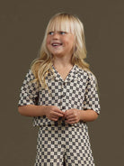 Smiling child in patterned outfit, hands clasped, against a neutral background.