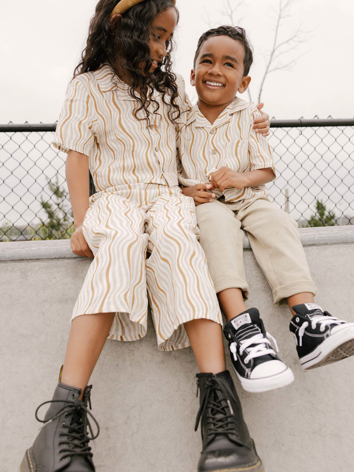 Two kids seated on a ledge, smiling. The girl wears a striped outfit and black boots; the boy wears a striped shirt and sneakers.