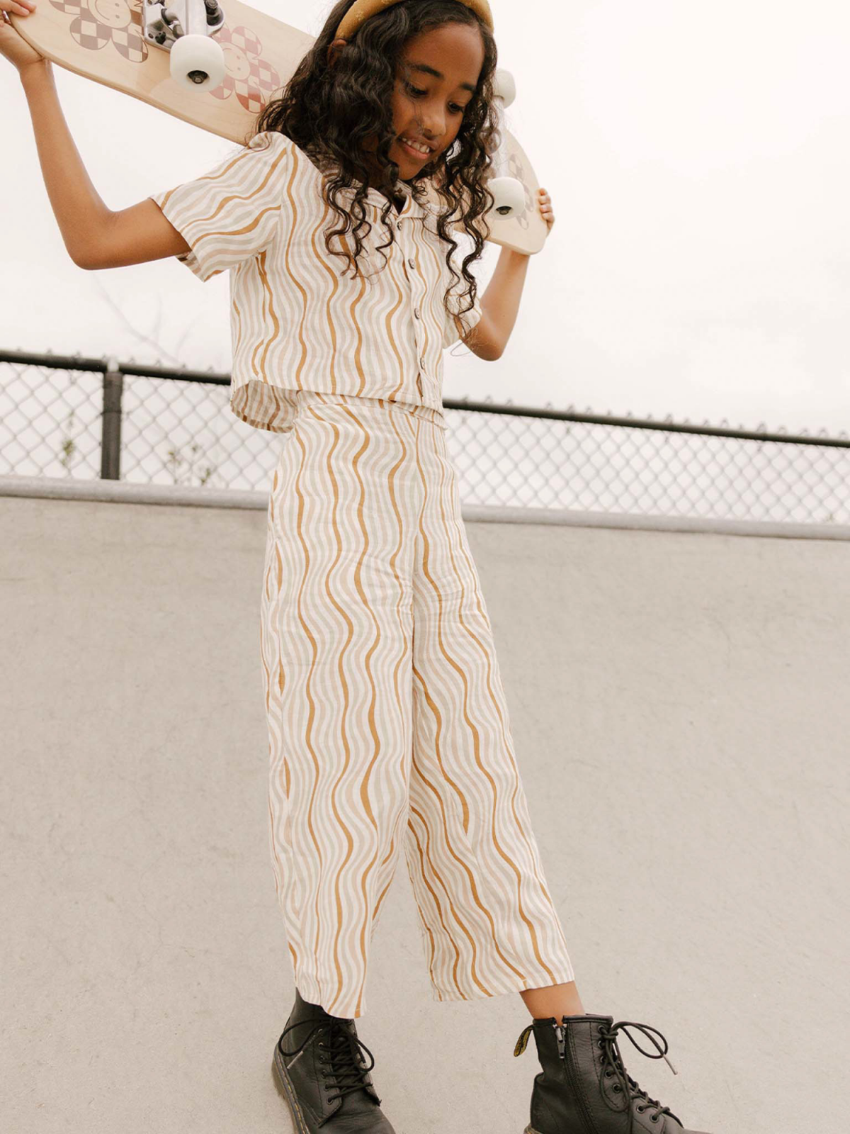 Girl with curly hair smiles while holding a skateboard, wearing a patterned outfit and boots.