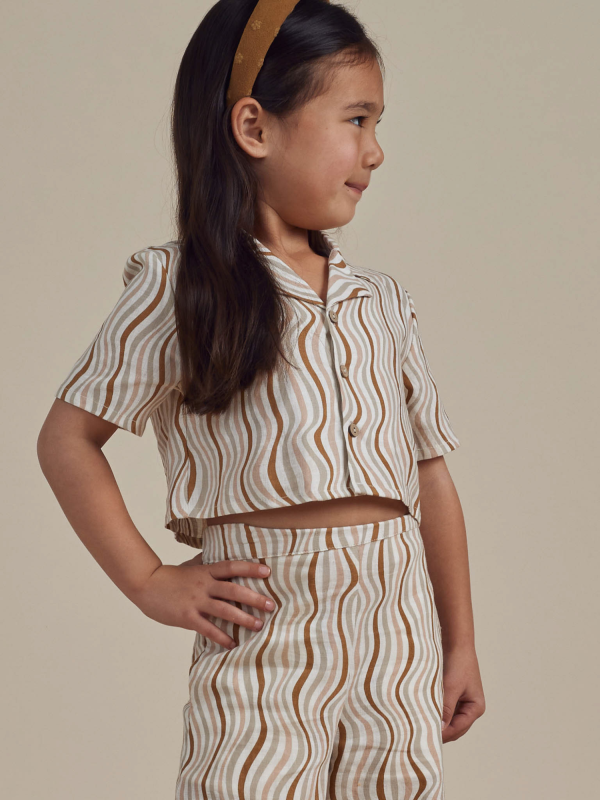 Girl in a striped two-piece outfit, smiling confidently with hands on hips, against a neutral background.