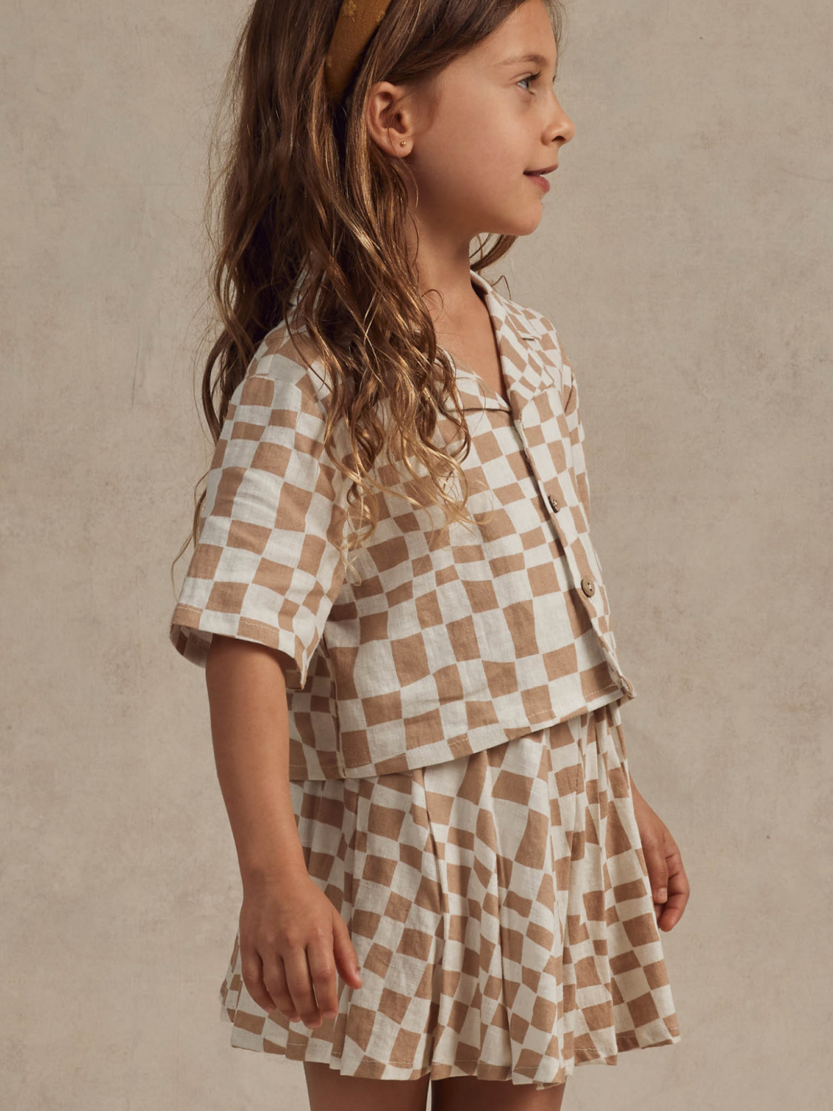Girl in a checkered outfit with wavy hair, smiling softly against a neutral background.