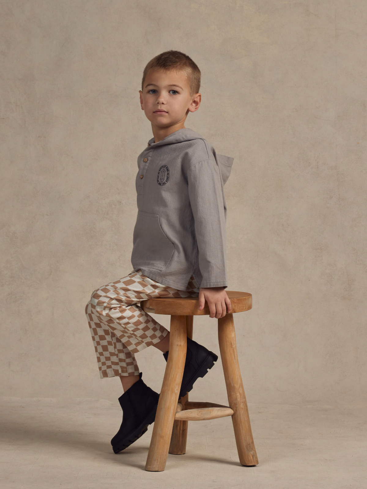 Child in a gray hoodie and checkered pants sits on a wooden stool against a neutral background.