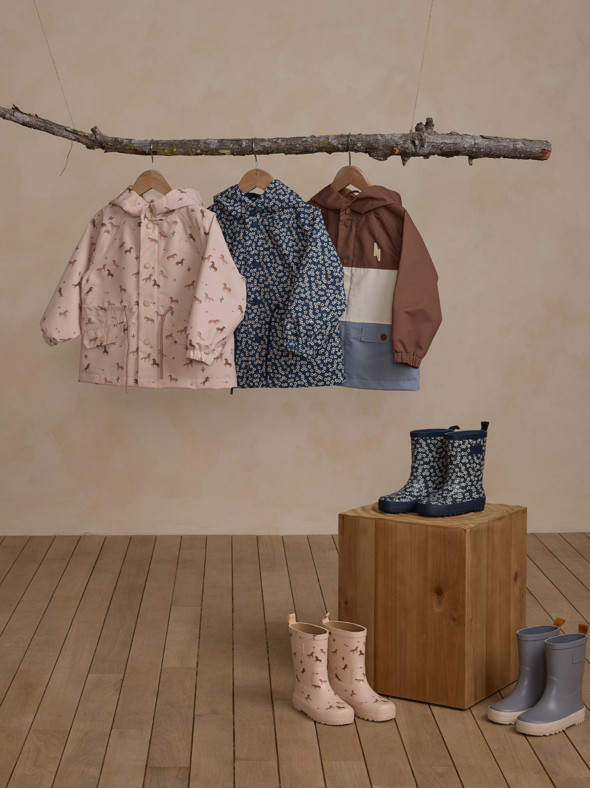 Three children's rain jackets hang from a branch above colorful rain boots on a wooden platform.