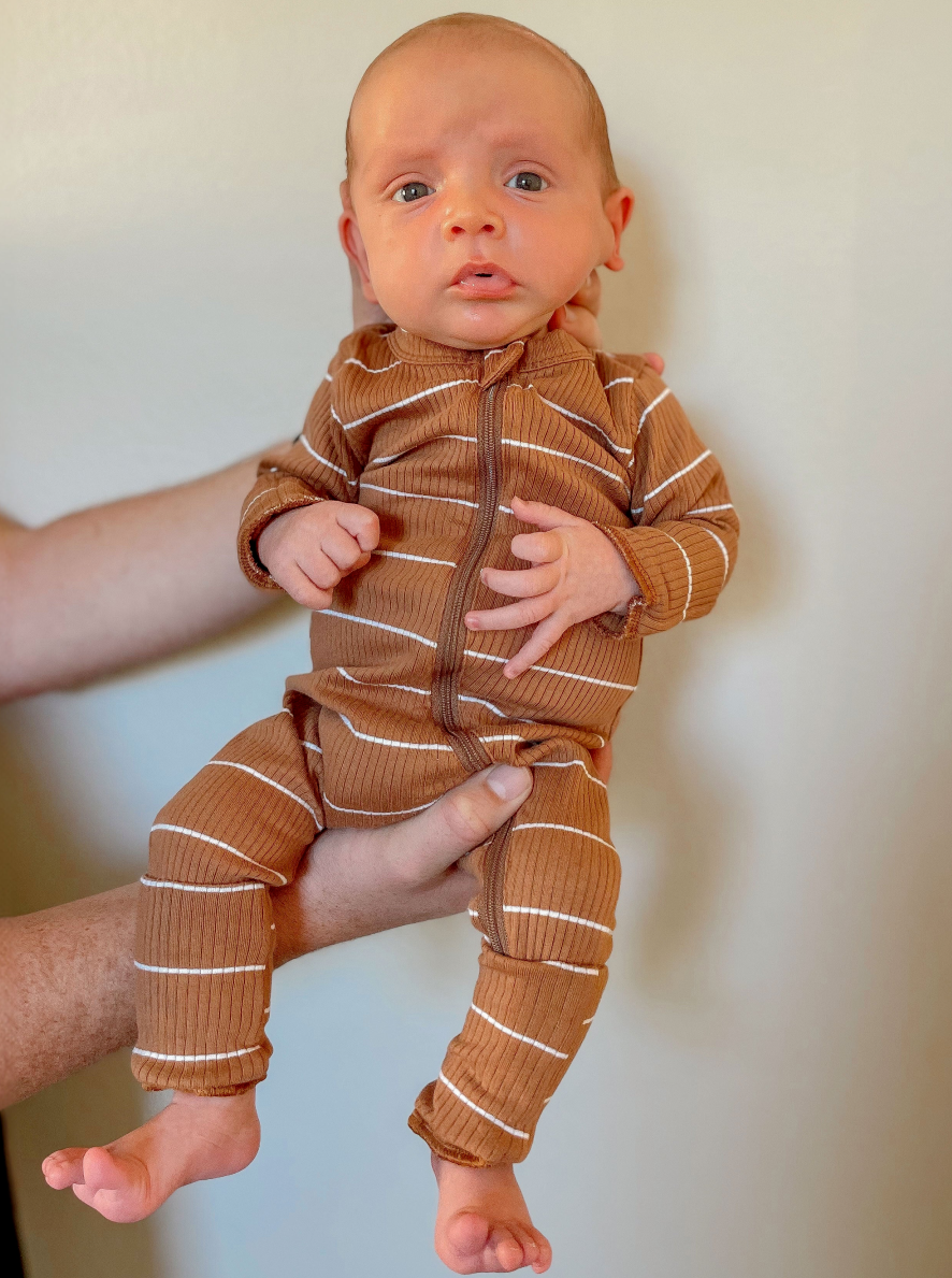 A baby in a brown striped onesie is held by a person against a light background.