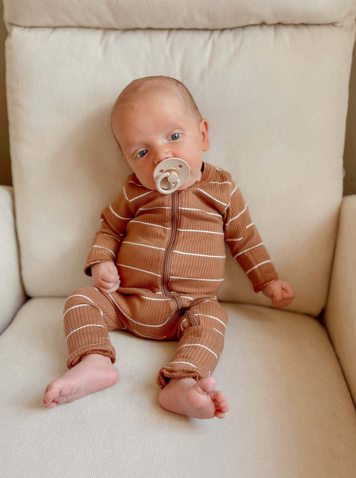 Baby in brown striped outfit sitting on a cream armchair, sucking on a pacifier.