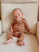 Baby in brown striped outfit sitting on a cream armchair, sucking on a pacifier.