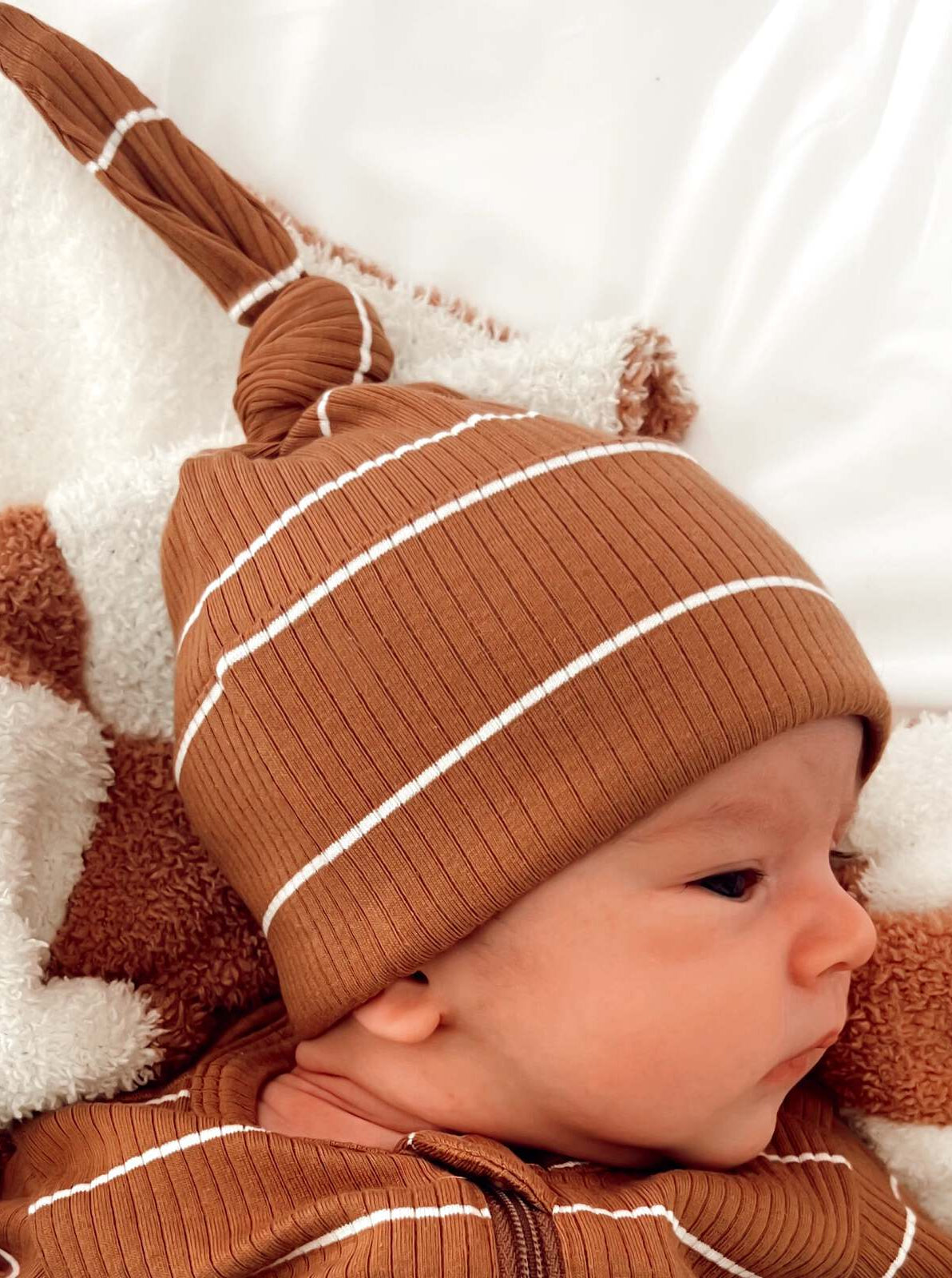 Baby wearing a brown ribbed hat with white stripes, resting on a soft blanket.