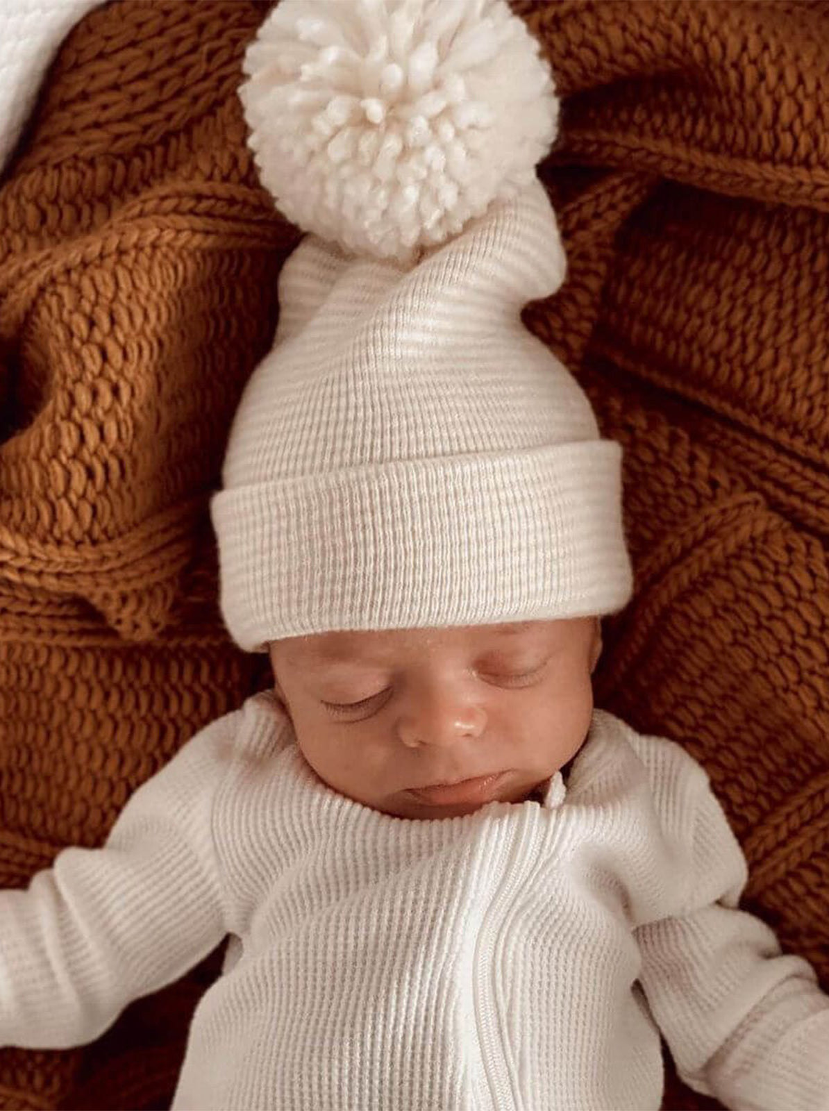 Newborn in a white outfit and cozy hat with pompom, resting on a textured brown blanket.