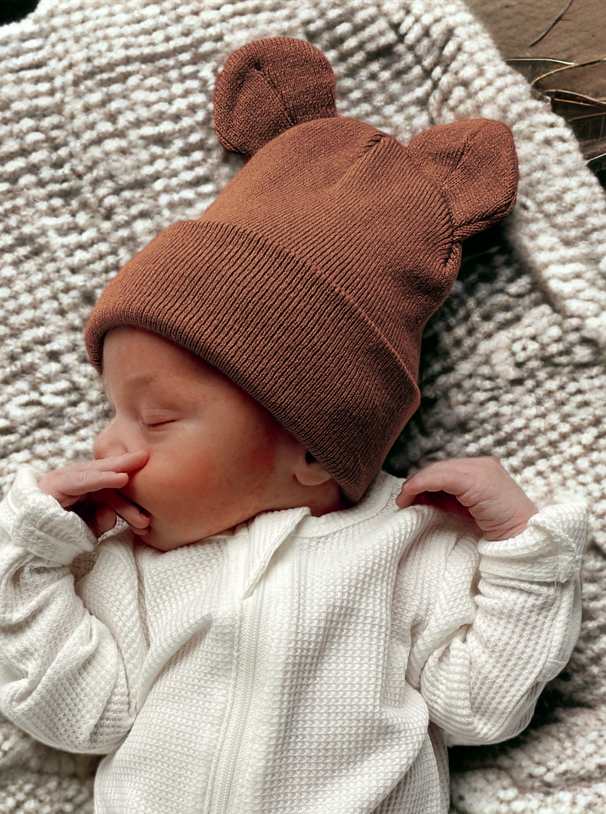 Sleepy baby in a brown bear hat, snuggled in white outfit on a cozy blanket.