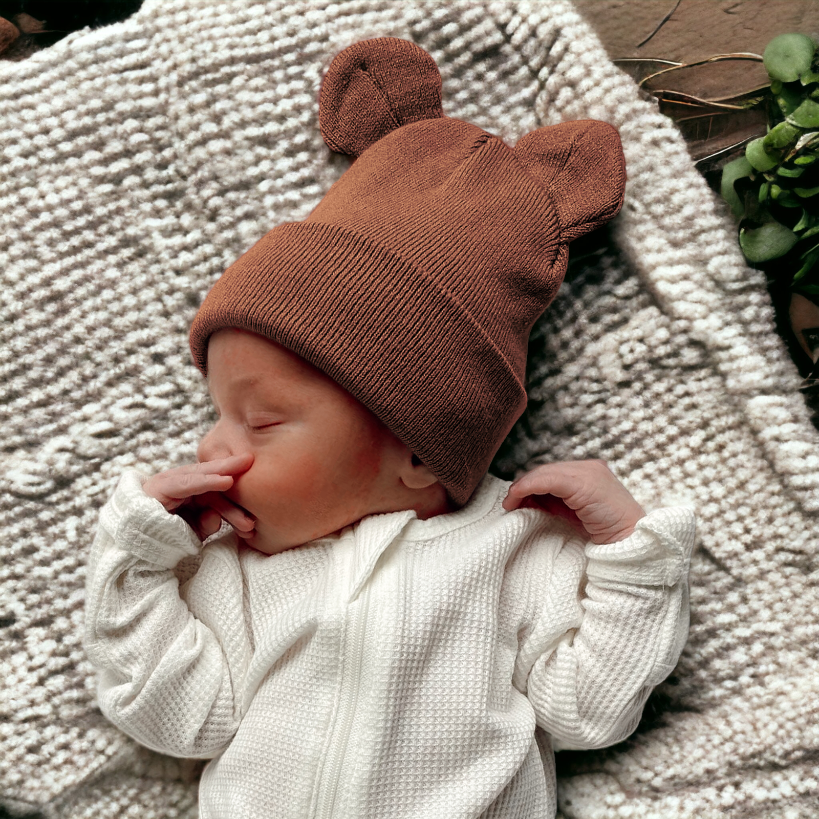 Sleepy baby in a brown bear hat, snuggled in white outfit on a cozy blanket.