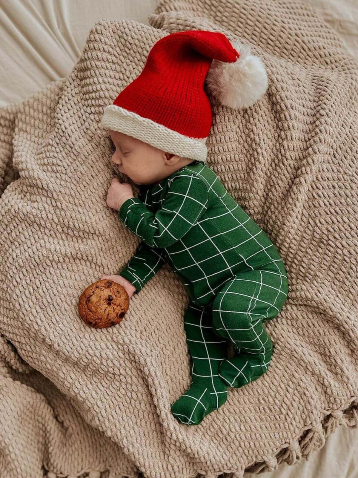 Sleeping baby in a festive red hat, wearing green pajamas, cuddling a cookie on a cozy blanket.