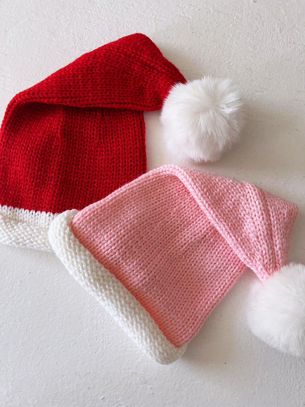 Red and pink knit holiday hats with white trim and pom-poms on a light background.