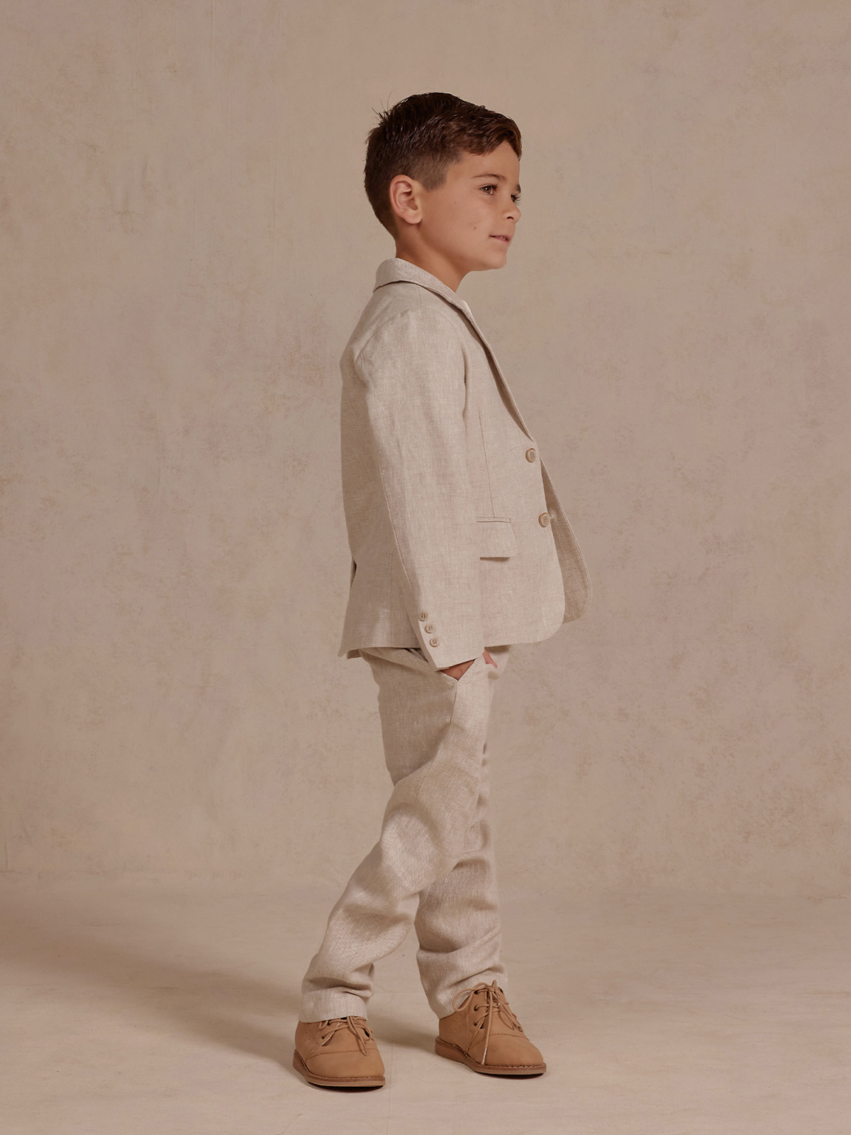 Child wearing a light beige suit with tan shoes, posing sideways against a neutral background.