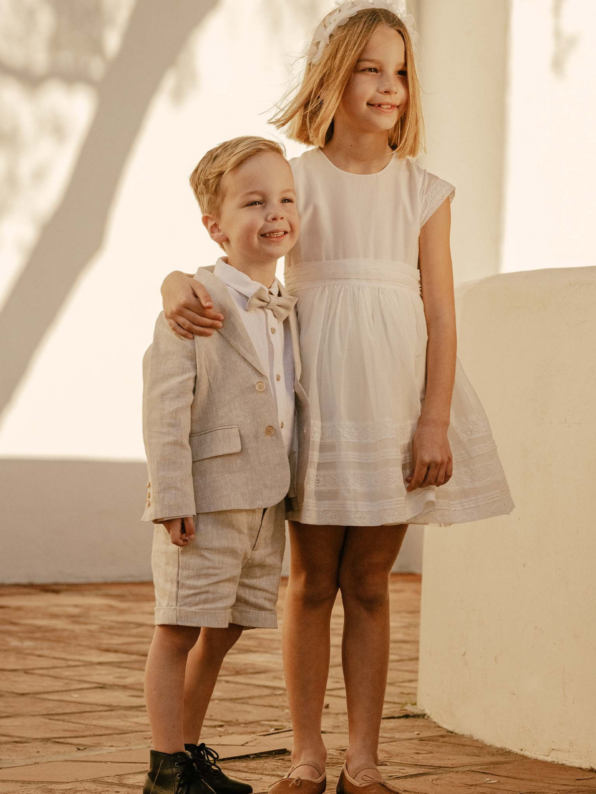 A boy in a light suit and a girl in a white dress stand together, smiling in a sunny outdoor setting.