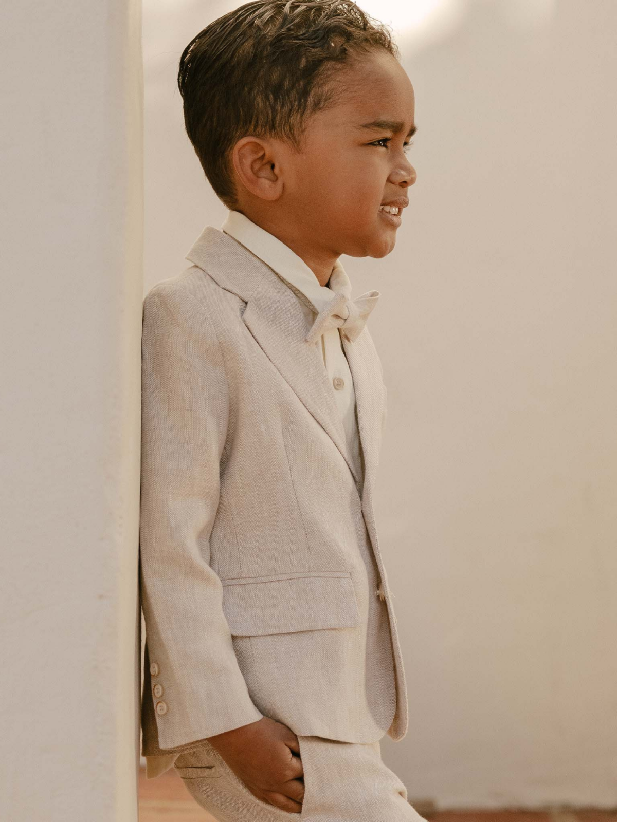 Young boy in a light suit and bow tie, leaning against a wall, smiling confidently.