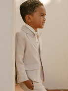 Young boy in a light suit and bow tie, leaning against a wall, smiling confidently.